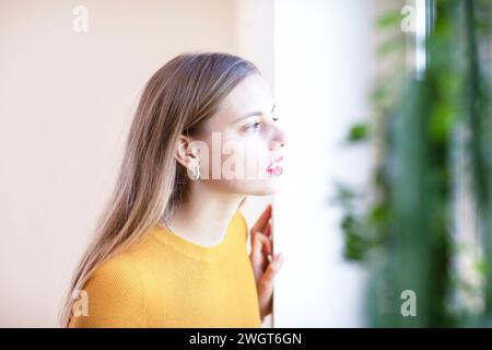 giovane donna con capelli biondi che guarda fuori dalla finestra Foto Stock