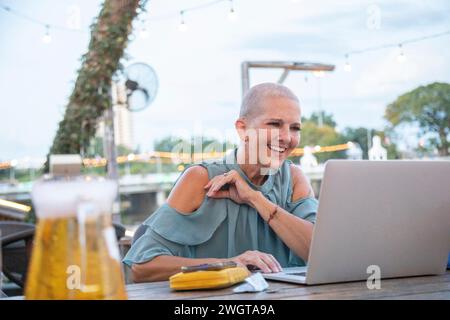 Una donna di mezza età siede in un ristorante sul fiume nel nord della Thailandia. Sta ridendo sul suo portatile. Foto Stock