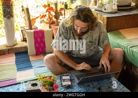 Young Man lavora al suo computer portatile e al telefono in un ristorante nel nord della Thailandia, è un nomade digitale che lavora in remoto. Foto Stock
