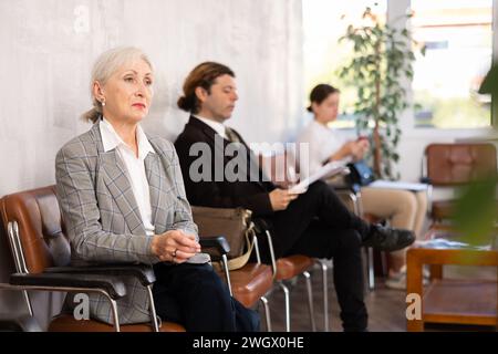 Una donna anziana e sicura di sé è seduta nella hall di una grande azienda, in attesa del suo turno per vedere uno specialista in ufficio Foto Stock