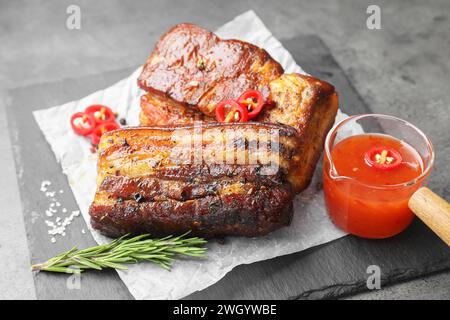 Pezzi di pancetta di maiale al forno serviti con salsa, rosmarino e peperoncino su tavola grigia Foto Stock