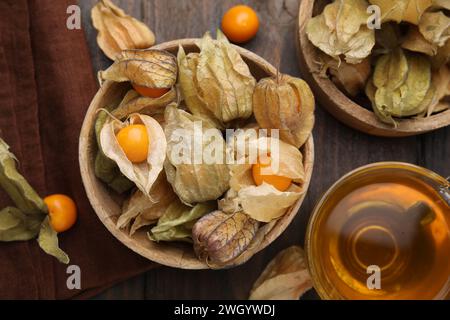 Frutti di physalis maturi con calici in ciotole e tazza di tè su tavola di legno, piatto Foto Stock