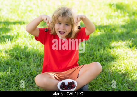 Il ragazzo si siede sull'erba e mangia ciliegia. Il ragazzo divertente mangia le ciliegie. Faccia eccitata. Ciliegia per bambini. Un bambino adolescente tiene le ciliegie. Cibo sano Foto Stock