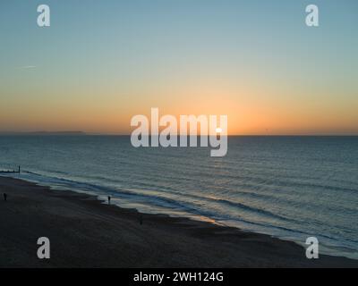 Il sole di gennaio è sorto sopra l'orizzonte sul mare calmo. Le persone si vedono in piedi da dietro, guardando il cielo azzurro e arancione che diventa più leggero Foto Stock