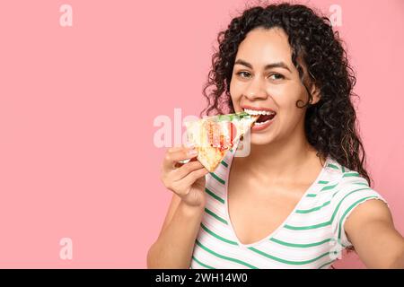 Bella giovane e felice donna afroamericana che mangia fetta di deliziosa pizza scattando selfie su sfondo rosa Foto Stock