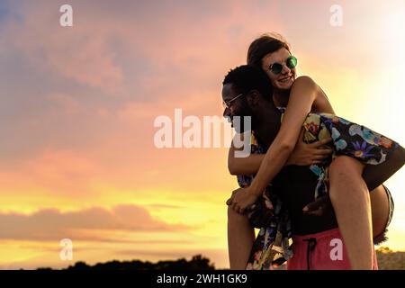 Una divertente coppia interrazziale si gode un giro in piggyback lungo la costa sotto il tramonto dorato Foto Stock