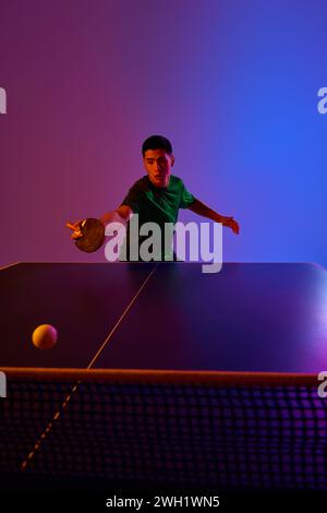 Tennistavolo concentrato in abbigliamento sportivo verde, tenendo d'occhio la palla, preparandosi per un tiro in fronte su sfondo gradiente. Neon. Gel dinamico Foto Stock