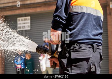 Un vigile del fuoco viene visto da dietro, maneggiando un tubo completamente innestato con un potente flusso d'acqua, probabilmente partecipando a un esercizio di allenamento o a un respondin Foto Stock