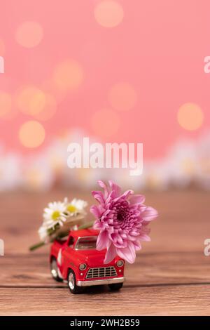 Camion rosso che trasporta un grande fiore sul retro del raccoglitore per consegnare il bouquet per San Valentino su sfondo rosa con campo di fiori sfocati Foto Stock