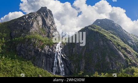 La luce del sole avvolge una maestosa cascata norvegese Valefossen che scende giù da aspre scogliere, circondata da vegetazione lussureggiante e vette spettacolari. Lofoten Foto Stock