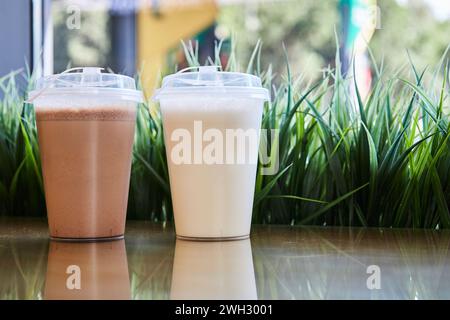 Due bicchieri di plastica trasparente con coperchi riempiti di frullati di cioccolato alla vaniglia sul tavolo, erba verde. Mockup vuoto, caffetteria con bevande di sottofondo. Portate via Foto Stock