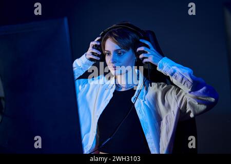 donna concentrata che indossa le cuffie e guarda il computer in una stanza illuminata dal blu, concetto di gioco cybersport Foto Stock