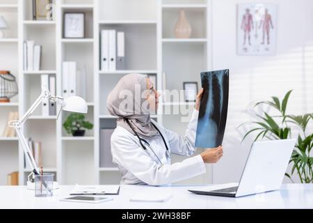 Donna musulmana concentrata medico con stetoscopio che controlla radiografia dei polmoni mentre si siede al gabinetto dell'ospedale con computer portatile. Polmonologo qualificato in uniforme medica bianca che fa la diagnosi per il paziente. Foto Stock