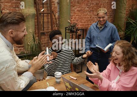 Schietto di giovani diversi che lavorano insieme al tavolo delle riunioni e applaudono per celebrare il successo Foto Stock