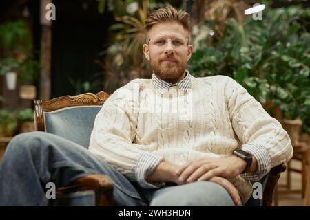 Ritratto di un giovane barbuto che indossa occhiali e maglione casual che si rilassa su una sedia vintage e guarda la fotocamera con sicurezza Foto Stock
