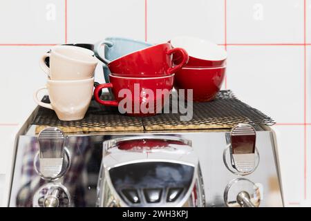 Set di tazze in ceramica colorata poggiano su una macchina da caffè in acciaio lucido di fronte a una parete bianca Foto Stock