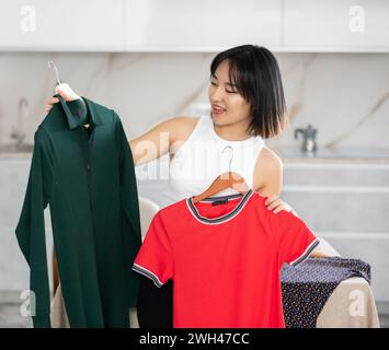 Felice giovane ragazza asiatica che cerca di scegliere un vestito in cucina Foto Stock