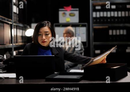 Investigatore privato asiatico che avvia un caso di indagine penale, confrontando i dettagli delle indagini criminali. Detective donna nell'ufficio archivio dei fascicoli pieno di cartelle criminologiche Foto Stock