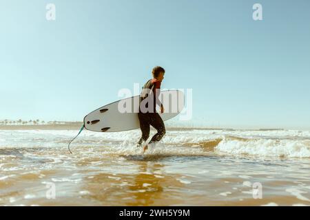 Vista laterale del surfista maschile in muta con la sua tavola da surf che entra in mare. Surf sull'oceano Foto Stock