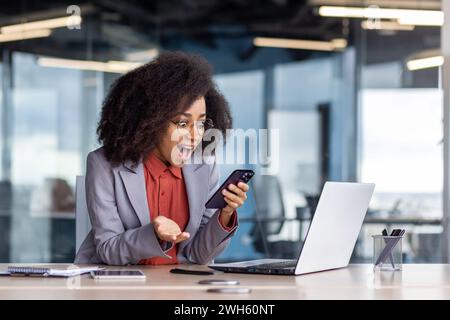 Giovane donna d'affari eccitata con capelli ricci si rallegra del suo successo mentre lavora in un ambiente d'ufficio contemporaneo. Foto Stock