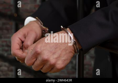 Uomo in tuta ammanettato, mani appoggiate su sbarre, sfondo sfocato di mattoni wal Foto Stock