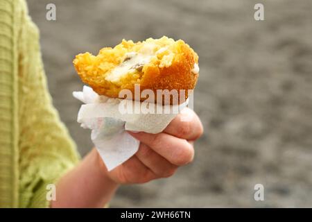 Mano della donna al mare che tiene in mano un caldo arancino (palle di riso fritte in profondità con carne). Tipico cibo di strada siciliano. Primo piano. Foto Stock