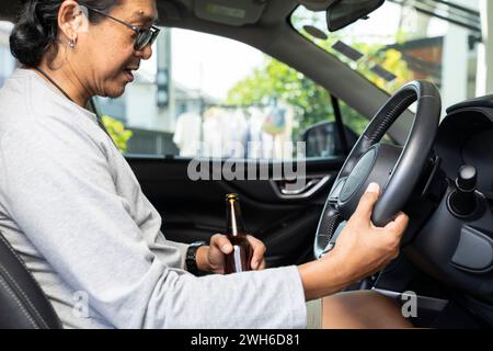 Un giovane asiatico ubriaco guida un'auto con una bottiglia di birra, un concetto di guida pericoloso. Foto Stock