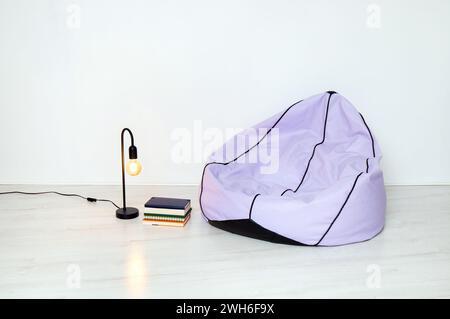 Concetto di casa minimalista. Sacchetto di fagioli viola, lampada in metallo nero e pila di libri in cestello metallico, pareti bianche e pavimento. Meno è di più. Foto Stock