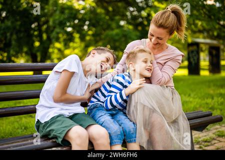 Madre felice è seduta con i suoi figli sulla panchina nel parco. Si stanno divertendo insieme. Foto Stock