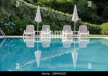Lettini e ombrelloni bianchi vuoti che si riflettono in una piscina blu Foto Stock