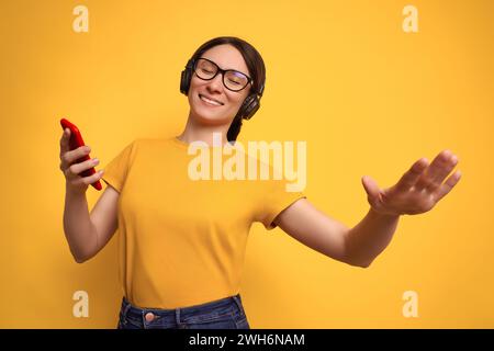 Foto in studio di una bella donna bruna con t-shirt gialla e cuffie wireless per ascoltare musica ad alto volume e ballare su sfondo giallo. Ragazza allegra goditi la playlist preferita con l'app per smartphone online Foto Stock