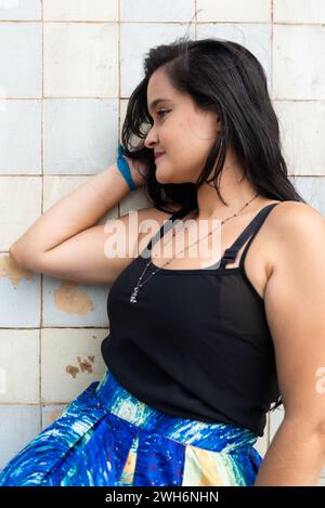 Ritratto di una bella donna, con lunghi capelli neri, appoggiata all'esterno su una vecchia parete piastrellata. Persona felice che viaggia. Foto Stock