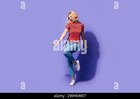 Foto a tutta lunghezza di una ragazza funky in un abbigliamento casual, che ama la musica pop su youtube premium utilizzando auricolari isolati su sfondo viola Foto Stock