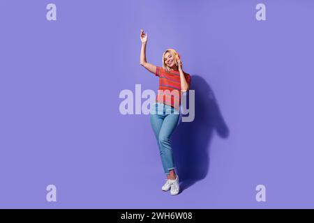 Foto a tutto il corpo di una donna bionda spensierata in jeans t shirt a righe che amano le moderne cuffie bluetooth isolate su sfondo di colore viola Foto Stock