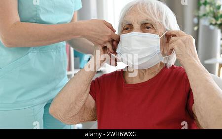 Infermiere aiuta la donna anziana a indossare la maschera protettiva medica durante il periodo di malattie virali e respiratorie Foto Stock