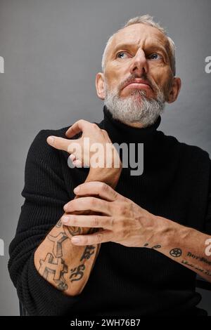 affascinante fashionista maschile maturo con barba grigia in un elegante dolcevita nero che posa e guarda lontano Foto Stock