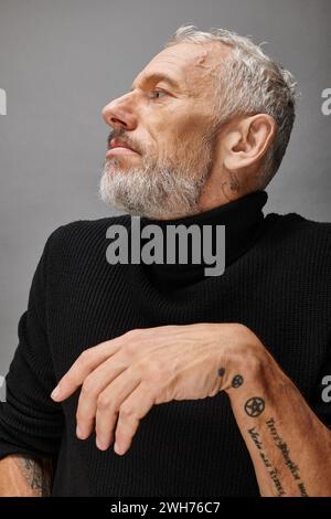 affascinante fashionista maschile maturo con barba grigia in un elegante dolcevita nero che posa e guarda lontano Foto Stock