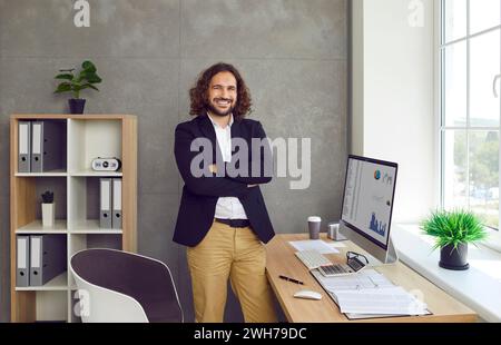 Uomo d'affari sicuro in ufficio vicino al suo posto di lavoro e che guarda la fotocamera. Foto Stock