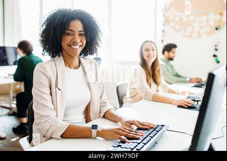 Un'allegra dipendente donna con capelli ricci irradia positività, mentre si impegna con il suo lavoro su un computer desktop in un ufficio open space, dimostrando un ambiente di lavoro produttivo e amichevole Foto Stock