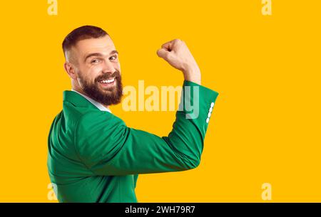 L'uomo felice in tuta verde sorride, si flette il braccio e mostra i suoi forti muscoli bicipali Foto Stock