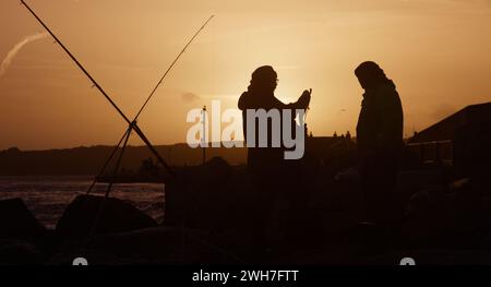 Sagoma di canne da pesca e pescatori che misurano Un branzino pescato al tramonto su Mudeford Quay, Avon Beach, Christchurch Regno Unito Foto Stock