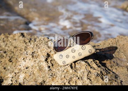 pietra naturale con facce divertenti e tristi dipinte su di essa e occhiali da sole sulla costa del mare. Ciao estate, sole, vacanze, emozioni positive, viaggi. Scegliere y Foto Stock