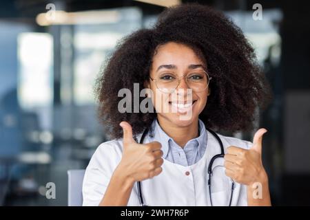 Una dottoressa con gli occhiali e un sorriso regala un pollice in alto, diffondendo vibrazioni divertenti durante un evento di cura della vista. Foto Stock