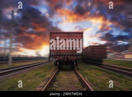 Treno merci in movimento con effetto sfocato contro il cielo del tramonto con nuvole. Trasporto ferroviario merci. Foto Stock