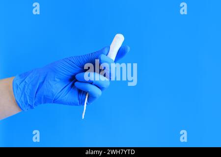 Mano femminile con tampone, sfondo blu. Una mano in un guanto medico in lattice blu contiene un tampone. Prodotto per l'igiene personale femminile. Pulire il tampo di cotone Foto Stock