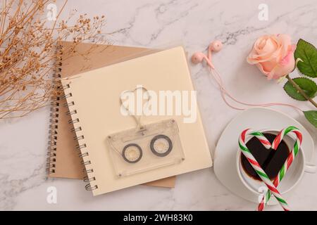 cassetta audio vintage trasparente con nastro magnetico a forma di cuore su una pagina vuota del libro, bastoncini di caramelle a forma di cuore sulla tazza del caffè, auricolari Foto Stock