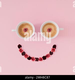 Disposizione dell'emoticon sorridente con tazze da caffè e fiori su sfondo rosa. Concetto di caffè minimale. Pensiero creativo positivo e idea di buon umore. Foto Stock