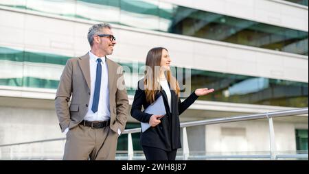 Due professionisti, un uomo anziano e una giovane donna in abbigliamento da lavoro, vengono catturati in uno scambio allegro mentre camminano, riflettendo un mix di discussione professionale e una cultura aziendale rilassata Foto Stock