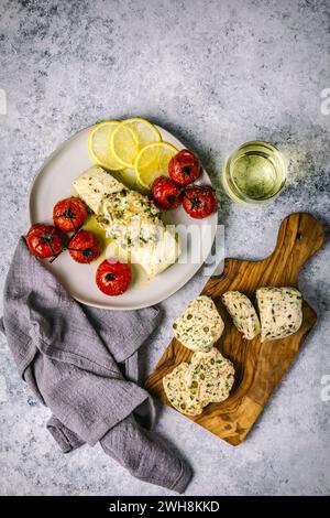 Colpo di testa con filetto di pesce bianco ricoperto di burro composto fuso su un piatto bianco con pomodori arrostiti, limoni a fette e vino bianco in un gambo Foto Stock