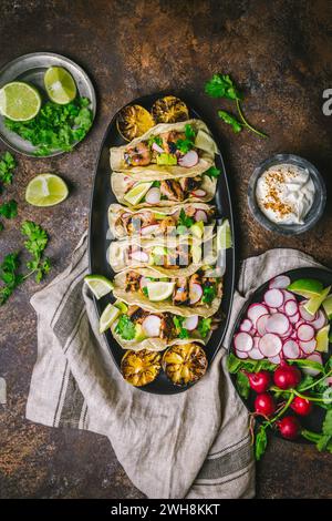 Disposizione di Tacos di pollo alla griglia su un piatto ovale scuro con ravanelli a fette, limes, coriandolo, limoni carbonizzati e panna acida Foto Stock